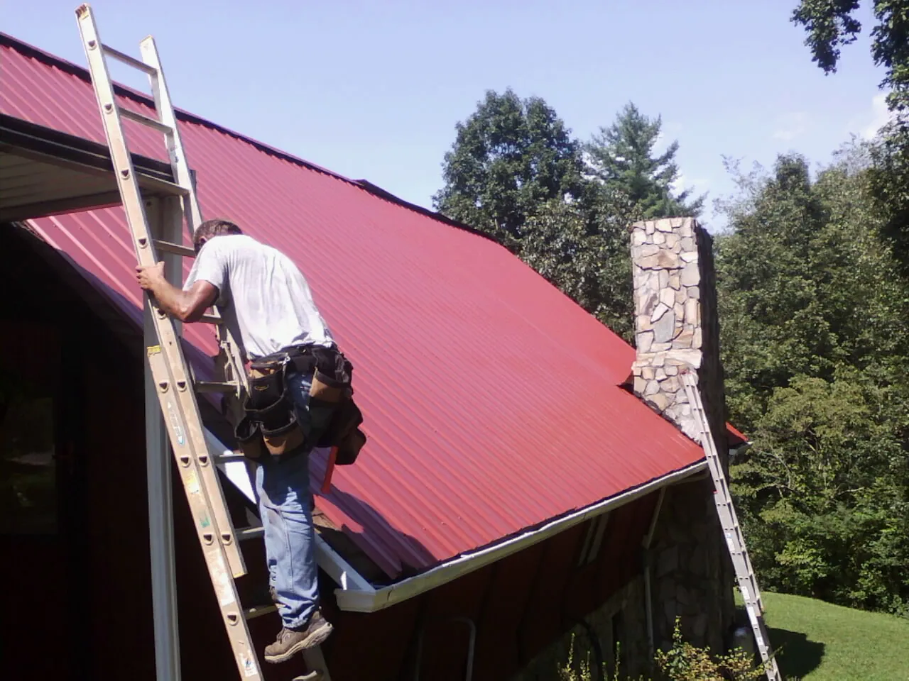 Roofer on a ladder beside a red metal roof and stone chimney — Soffit & Fascia Repair by Roofing Brothers in Saegertown